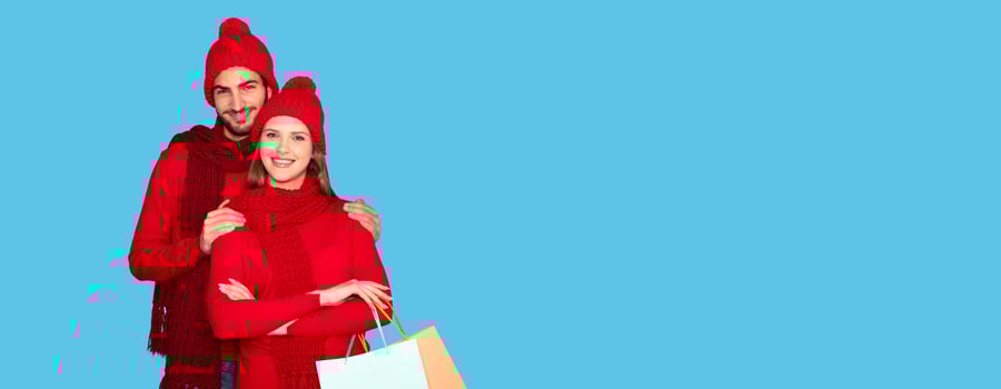 Happy Couple Wearing Winter Clothes Embracing And Holding Shopping Bags, Smiling Young Man And Woman Enjoying Seasonal Sales, Standing Isolated Over Blue Studio Background, Copy Space photo