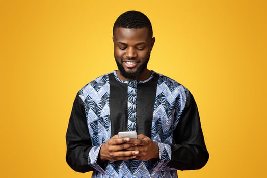 Smiling black guy using modern smartphone, wearing african t-shirt, pink studio background. African american young man in traditional costume chatting with friends on mobile phone, copy space photo