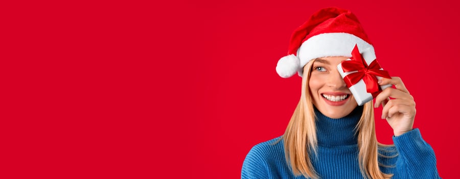 Happy lady wearing Santa hat, holding mini gift box and covering eye, looking aside at free space over red studio background. Christmas, New Year gifts and celebration photo