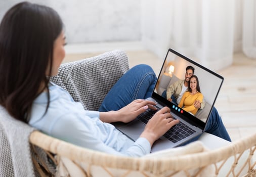 A woman engaged in a call on her laptop, smiling at a couple on the screen. She is seated in a cozy chair, with a bright, airy room in the background. photo
