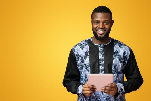 Positive black guy using digital tablet, wearing african t-shirt, pink studio background. African american young man in traditional costume checking social media on newest pad, copy space photo