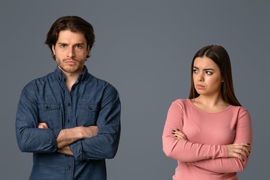 Young woman looking with sorrow at her offended husband after their argue, couple standing with folded arms over grey background, empty space photo