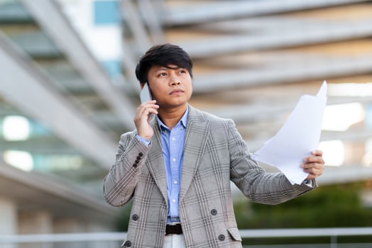 A young Asian man stands outdoors, wearing a gray blazer and blue shirt, engaged in a phone conversation while holding important papers, showcasing a moment of business urgency. photo