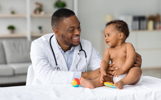 Child's Healthcare Concept. Portrait Of Smiling Black Doctor Making Check Up For Little Infant Baby Boy, Cute Toddler Child In Diaper Having Appointment At Pediatrician's Office, Free Space photo
