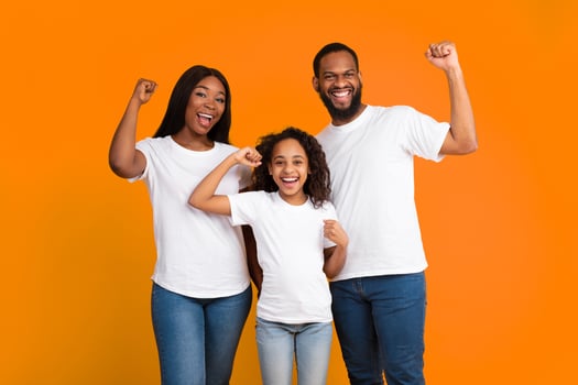 Yes. Portrait of excited young black family of three people shaking clenched fists, making winner gesture. Happy African American man, woman and girl celebrating victory together, yellow orange studio photo
