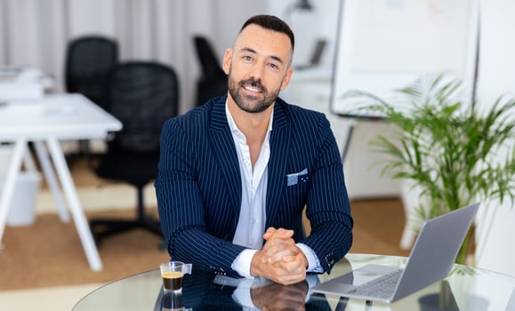 A man in a striped suit sits confidently at a glass desk in a contemporary office. He has a cup of coffee beside his laptop, ready for a productive workday ahead. photo