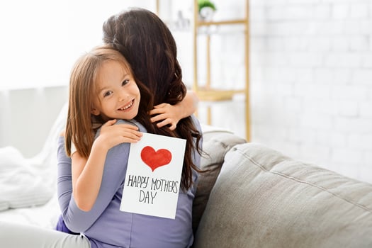 Smiling little daughter with Happy Mother's Day gift card hugging her mom, celebrating International Mother's Day together at home photo