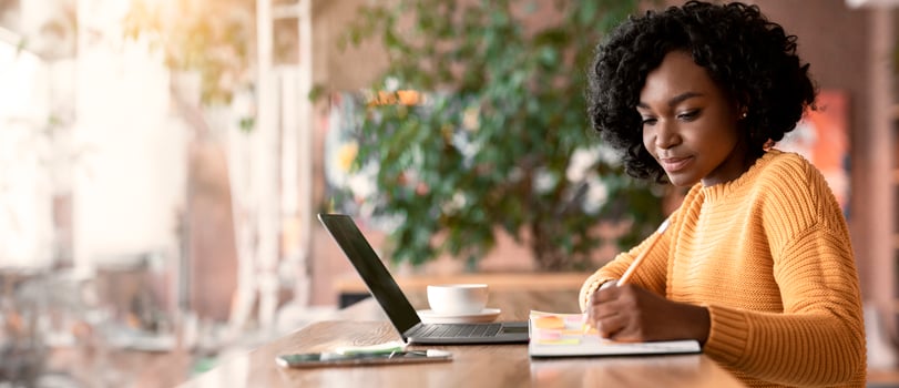 Motivated afro young woman looking for new job online, using laptop at cafe, taking notes, side view, copy space photo