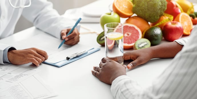 Detox concept. Afro woman patient of nutritionist drinking lemon water while doctor taking notes, close up photo