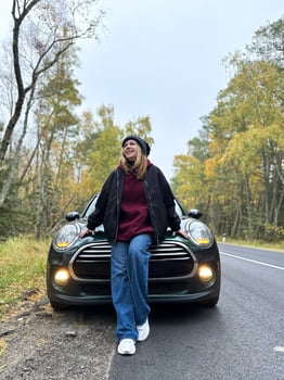 A young woman leans against the hood of her car, parked on a quiet autumn road surrounded by trees with golden foliage. Dressed in a black jacket, burgundy hoodie, and jeans, she gazes up with a smile photo