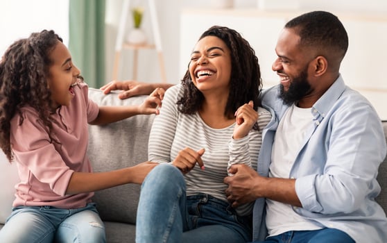 Portrait of cheerful African American man, woman and girl playing and having fun at home together, daughter and husband tickling laughing wife sitting on the couch in living room photo