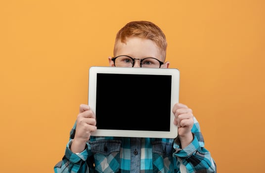 Ginger boy in eyeglasses hiding behind digital tablet with empty screen over yellow studio background. Cute redhead schoolboy showing pad, mockup, advertisement for cute application photo