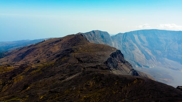 An aerial view of Mount Tambora's peak reveals a breathtaking landscape under a blue sky, as the sun rises, casting golden hues over the stunning terrain. photo