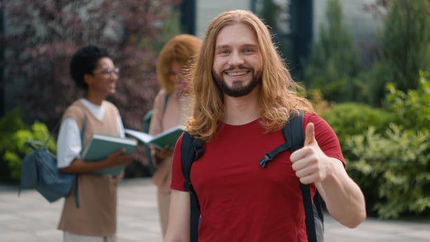 Happy Caucasian student guy man male schoolboy in city outdoors university college campus showing thumbs up to camera smile enjoy education study knowledge generation Z students talking on background photo