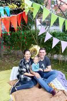 Smiling mom and dad with little girl sitting on a blanket in the garden near colorful flags photo