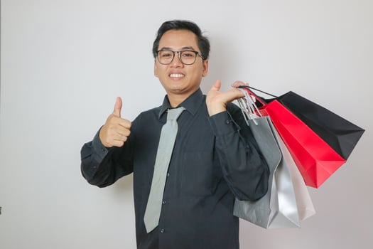 smiling and funny young Asian man in his 30s in black t-shirt standing holding shopping bags with groceries after shopping while showing thumbs up isolated on pastel white background studio portrait. photo