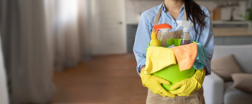 The individual in gloves carries a vibrant green container filled with cleaning tools and supplies while preparing to clean a well-lit living space with soft furnishings. photo