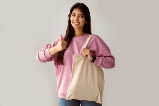 Zero Waste. Millennial Woman Showing Blank Eco Bag And Gesturing Thumbs Up Posing Standing On White Studio Background, Smiling To Camera. I Like Ecology Friendly Lifestyle, No More Plastic Concept photo