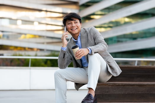 A young man, dressed in a stylish blazer and light pants, sits on a bench, smiling as he talks on his phone and enjoys a cup of coffee in an urban environment surrounded by contemporary architecture. photo