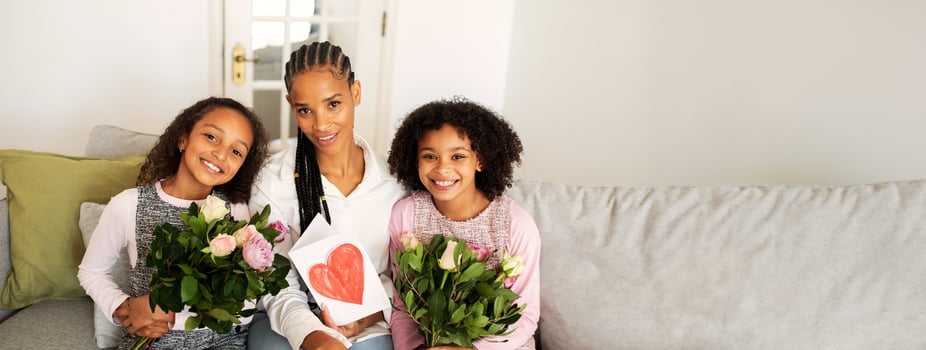 A black mother smiles proudly with her two daughters, each holding beautiful bouquets of flowers. They are seated comfortably on a couch, creating a joyful atmosphere in their living room. photo