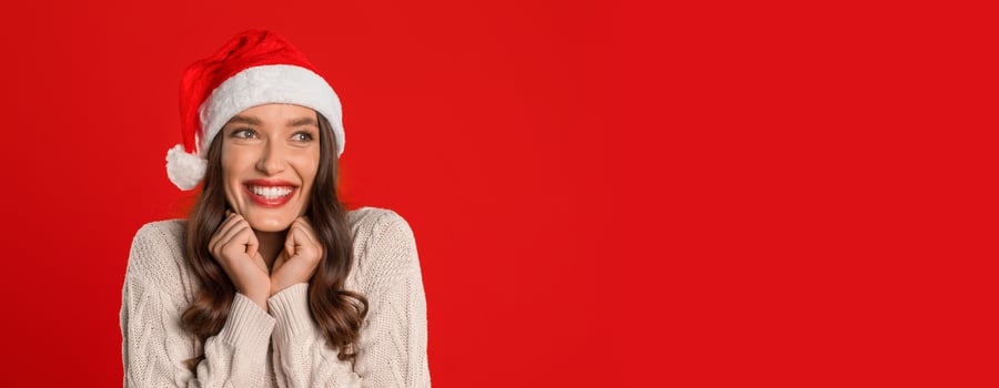 Smiling European lady in Santa hat looks aside anticipating festivities, against red studio backdrop, embodying the joy of Christmas and New Year season. Panorama, copy space photo