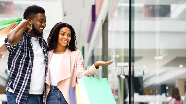 Discount Prices. Smiling Black Couple Shopping In Mall Together, Looking At Showcase With Interest, African Spouses Walking In Department Store With Bright Shopper Bags, Enjoying Seasonal Sales photo