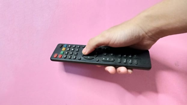 Close up of a hand holding a black TV remote against a pink background. Concept of entertainment, technology, and control. photo