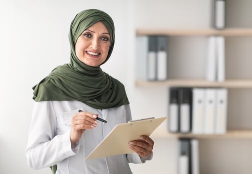 Happy Muslim Female Doctor Posing Smiling To Camera Holding Medical Case Record Standing In Modern Clinic Office Indoors. Portrait Of Mature Middle-Eastern Professional Therapist Lady Wearing Hijab photo