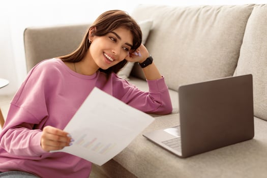 Portrait of positive young female manager working, holding paper or financial document, using laptop and cheking financial report. Woman sitting near the sofa with pc at home office, selective focus photo