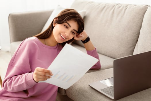 Paperwork Concept. Portrait of smiling young woman working at home office, holding and reading paper or financial document, sitting near the couch with laptop computer in living room photo