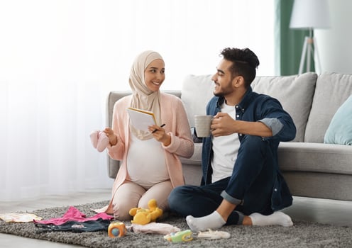 Happy pregnant muslim couple getting ready for childbirth, making checklist of necessities while sitting together on floor in living room with baby clothes around, islamic spouses relaxing at home photo