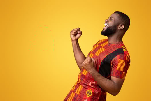 Emotional black young man in african costume celebrating success over yellow background photo