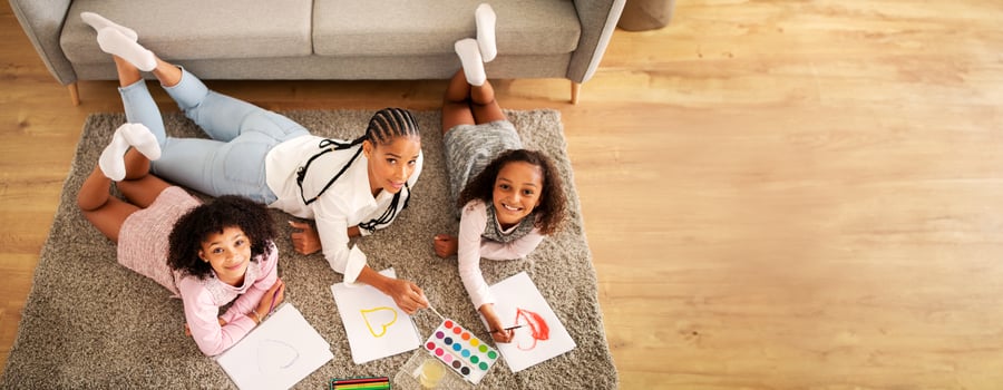 Black mother and children are lying on a soft rug, actively coloring and painting. A caregiver is assisting them, surrounded by art supplies and colorful drawings in a cozy living room setting. photo
