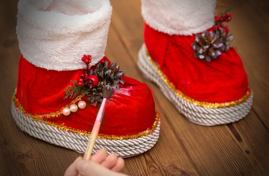 A girl with a paintbrush paints the red boots of santa claus. Decorations for the new year. Handmade, wooden background. Celebratory photo