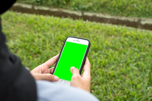A joyful woman in a black hijab and blue shirt smiles brightly while holding a smartphone, standing at the edge of a beautiful park sidewalk photo