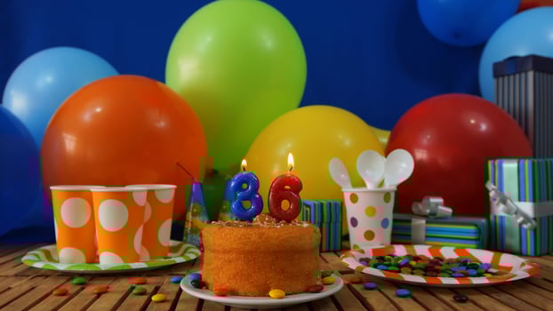 Birthday cake 36 on rustic wooden table with background of colorful balloons, gifts, plastic cups and plastic plate with candies and blue wall in the background photo