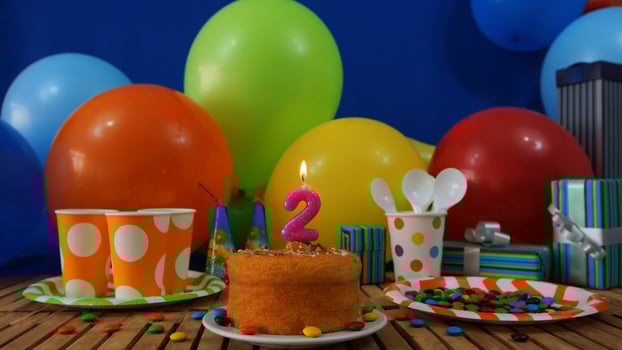 Birthday cake 2 on rustic wooden table with background of colorful balloons, gifts, plastic cups and plastic plate with candies and blue wall in the background photo