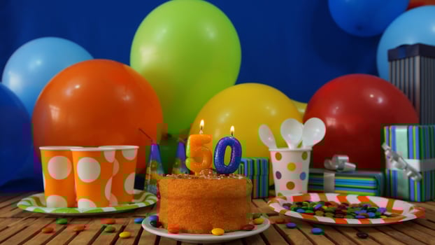 Birthday cake 50 on rustic wooden table with background of colorful balloons, gifts, plastic cups and plastic plate with candies and blue wall in the background photo