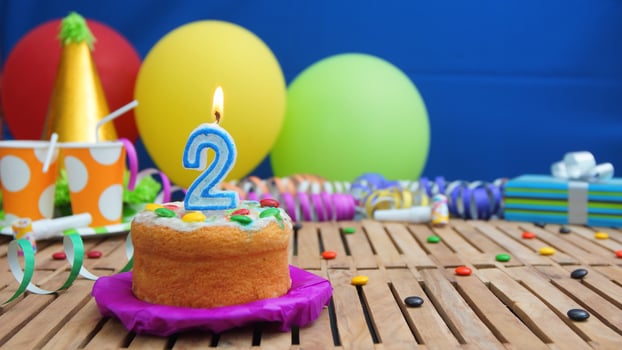 Birthday cake 2 with candles on rustic wooden table with background of colorful balloons, gifts, plastic cups and candies with blue wall in the background. Focus is on cake photo