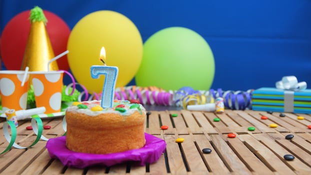 Birthday cake 7 with candles on rustic wooden table with background of colorful balloons, gifts, plastic cups and candies with blue wall in the background. Focus is on cake photo
