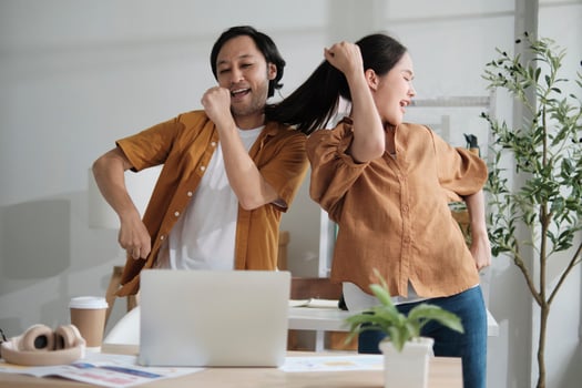 Happy working in casual small business office. Two Asian startup company colleagues enjoy dancing and singing songs together, relaxing from freelance jobs, humor, and funny coworker team in workplace. photo