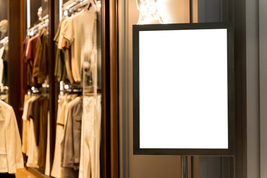White billboard in brightly lit clothing store mockup photography. Racks of apparel. Blank template advertising inside. photo