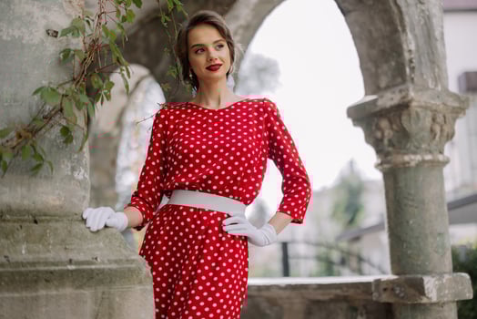 Elegant woman posing in red polka dot dress and white gloves near old building photo