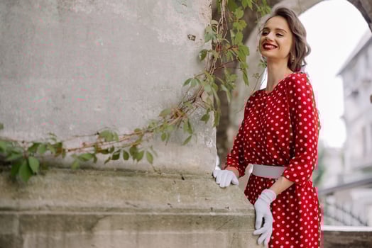 Elegant smiling woman wearing red polka dot dress and white gloves posing near old wall with climbing plant photo