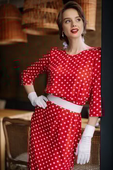 Elegant woman wearing red polka dot dress and white gloves posing in vintage interior photo