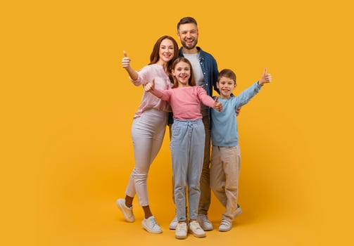 Happy family consisting of a mother, father, son, and daughter stands close together, all giving a thumbs up sign. They are posing against a vivid yellow backdrop photo