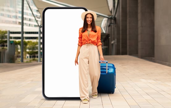 A young woman in an orange top and beige wide-leg pants walks confidently with a blue suitcase in an urban area, being framed by a large smartphone screen that symbolizes travel. photo