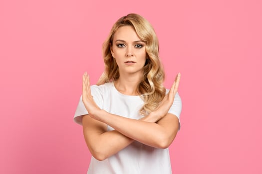 A woman with wavy blonde hair stands confidently, arms crossed in a stop gesture. She is wearing a white top and has a serious expression on her face, set against a vibrant pink backdrop. photo