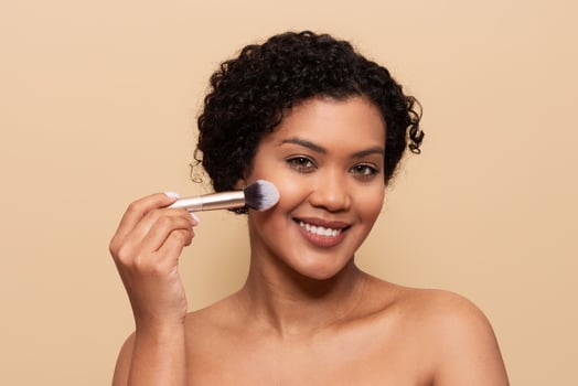 A young woman is smiling while applying blush to her cheeks with a makeup brush. She has curly hair and is against a plain background, emphasizing her joyful expression and beauty routine. photo