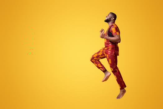 Happy barefoot black man in african costume jumping in air over yellow studio background, side view photo